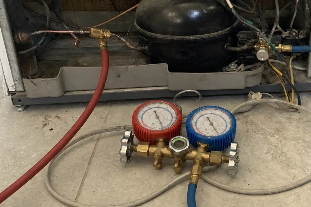 A red and blue manifold gauge set standing on a tiled floor with its hoses run up to the copper lines of an opened appliance base, the black compressor body behind them